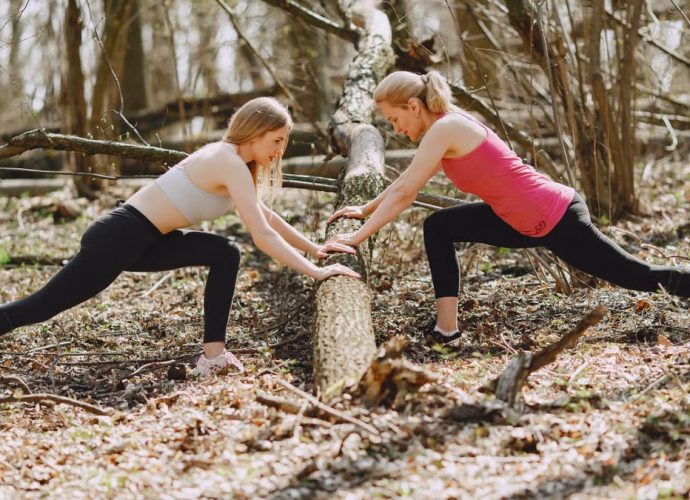 Side view of young female athletes in activewear stretching legs together using fallen tree during fitness workout in spring forest adhering to healthy lifestyle